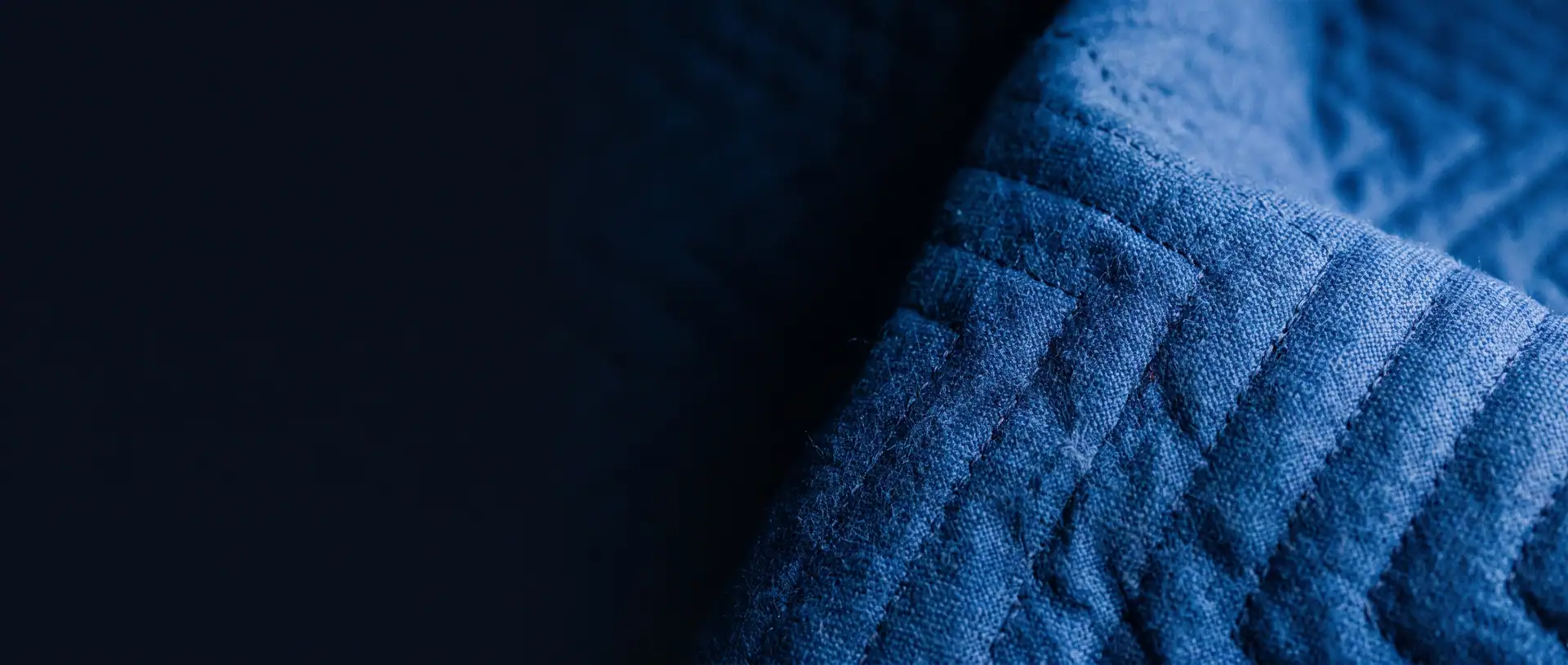Close view of a royal blue quilted moving blanket with zigzag stitching, lit from the right and fading into dark navy on the left